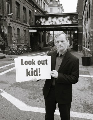 Theater Hora - Body Dylan. Ein Mann mit Hut und dunkler Sonnenbrille hält ein Schild hoch. Darauf steht: Neil Young.